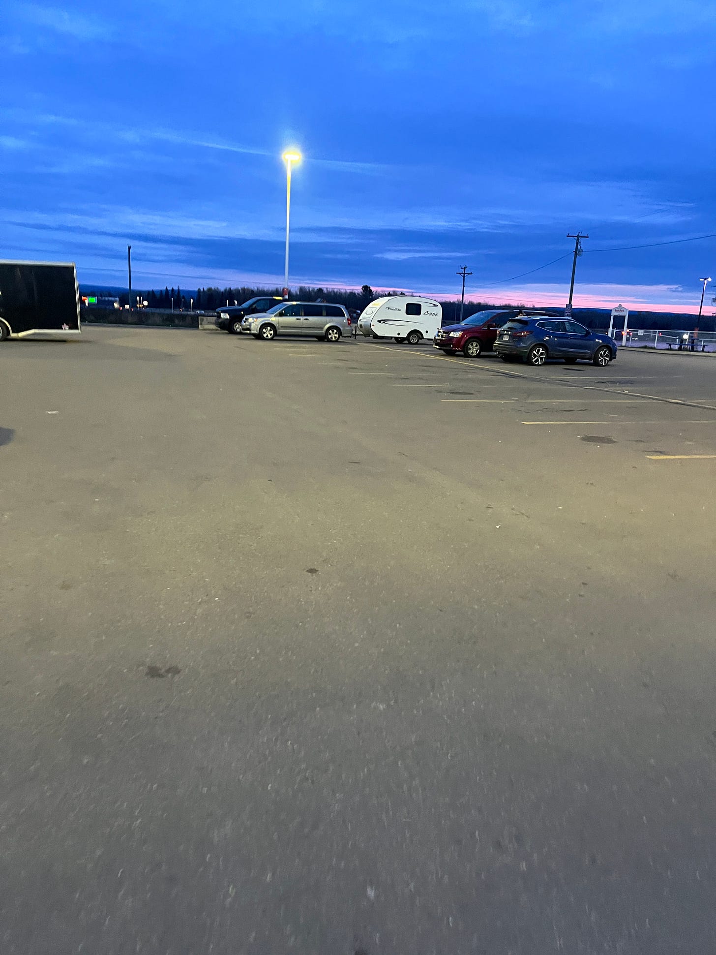 A long distance photo of my silver minivan parked with our camper trailer attached behind. It's in a large parking lot with a pretty purple and pink evening sky