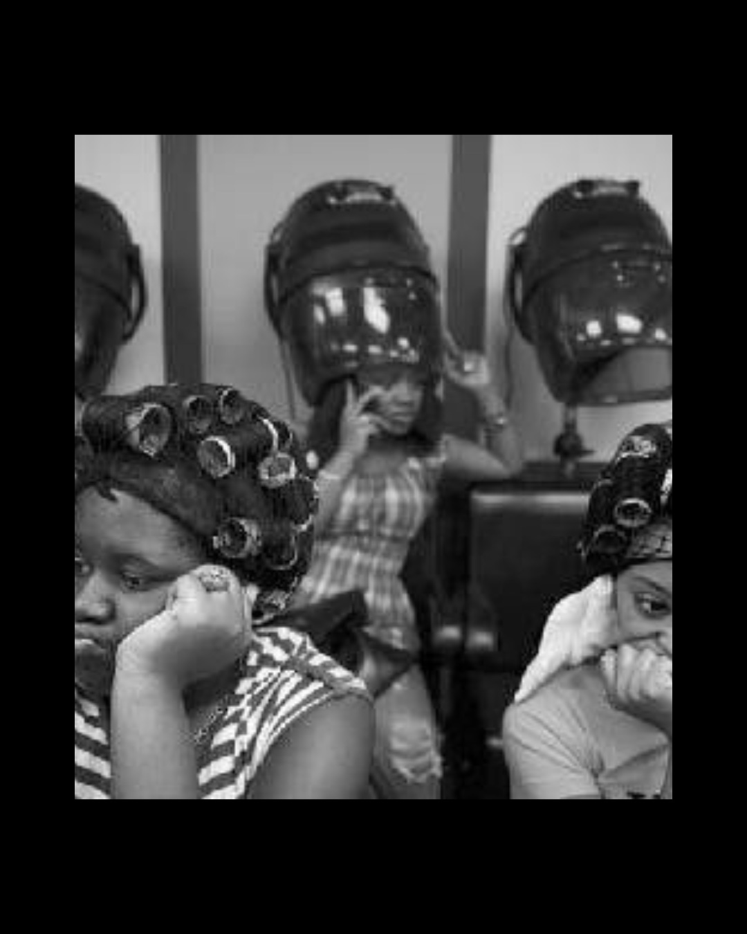 Black girls sit quietly beneath hooded hair dryers in a beauty salon.