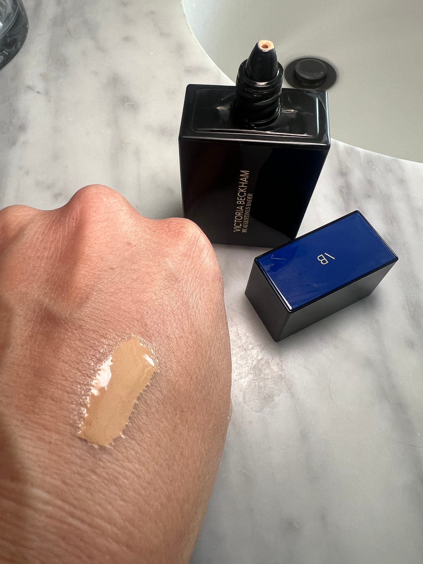 Foundation shade (5 Light) swatched on the back of a hand, showing the product's blendable texture, with the Victoria Beckham TFC8 bottle resting in the background.