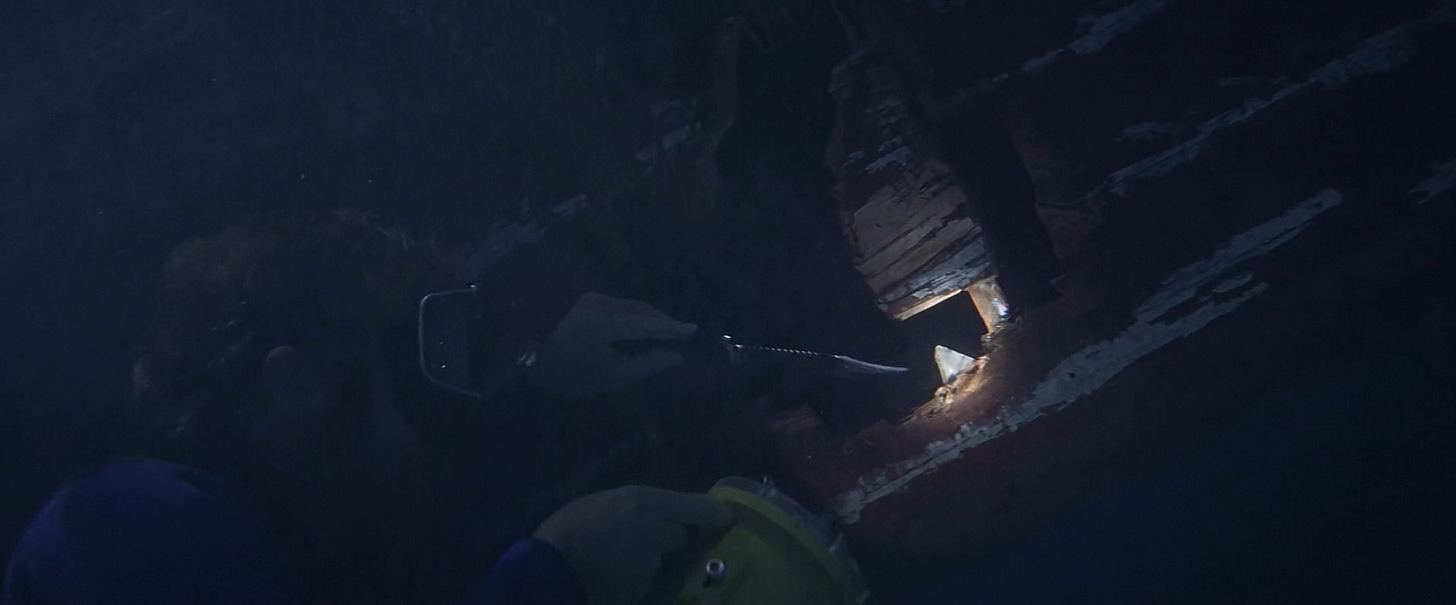 A frame from Jaws, underwater, showing a skin-diving Matt Hooper using a dive knife to dig a huge tooth out of the ragged edge of a hole in the side of motor boat.
