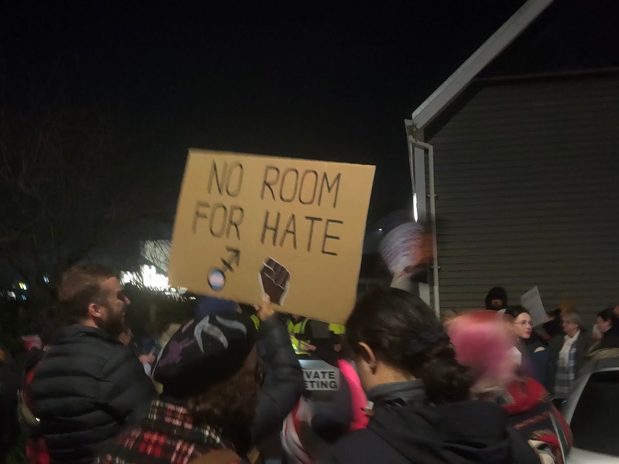 **Alt text:** A nighttime protest scene with a crowd of people standing outside near a building. In the center, someone holds a cardboard sign that reads “NO ROOM FOR HATE” with a raised fist symbol. The crowd is bundled in jackets, and several people appear blurred as they move through the scene. **Alt text:** A nighttime protest scene with a crowd of people standing outside near a building. In the center, someone holds a cardboard sign that reads “NO ROOM FOR HATE” with a raised fist symbol. The crowd is bundled in jackets, and several people appear blurred as they move through the scene.