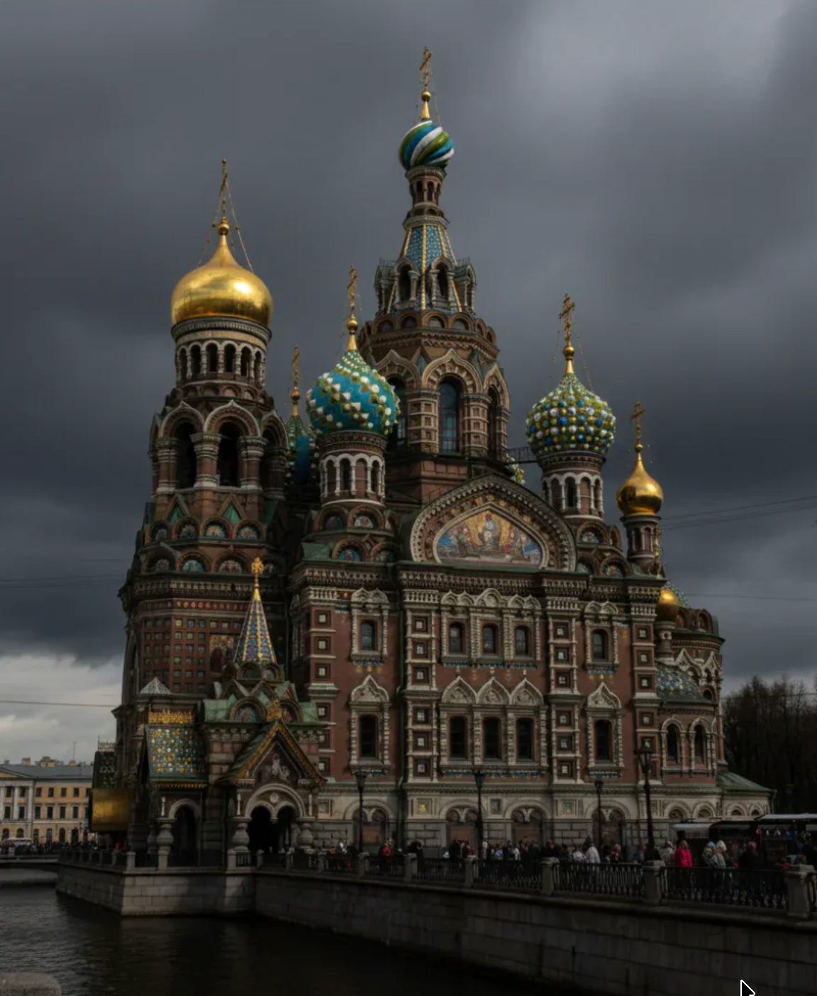 The Church of the Savior on Spilled Blood with its distinctive striped onion domes in gold, turquoise, and green against a heavy grey sky, reflecting in the dark waters of the canal.
