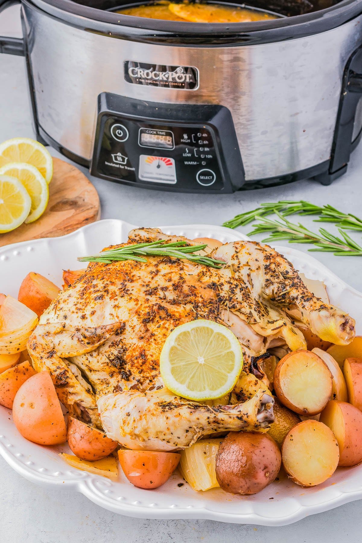 A cooked whole chicken garnished with rosemary and lemon slices rests on a platter surrounded by roasted potatoes, with a slow cooker visible in the background.
