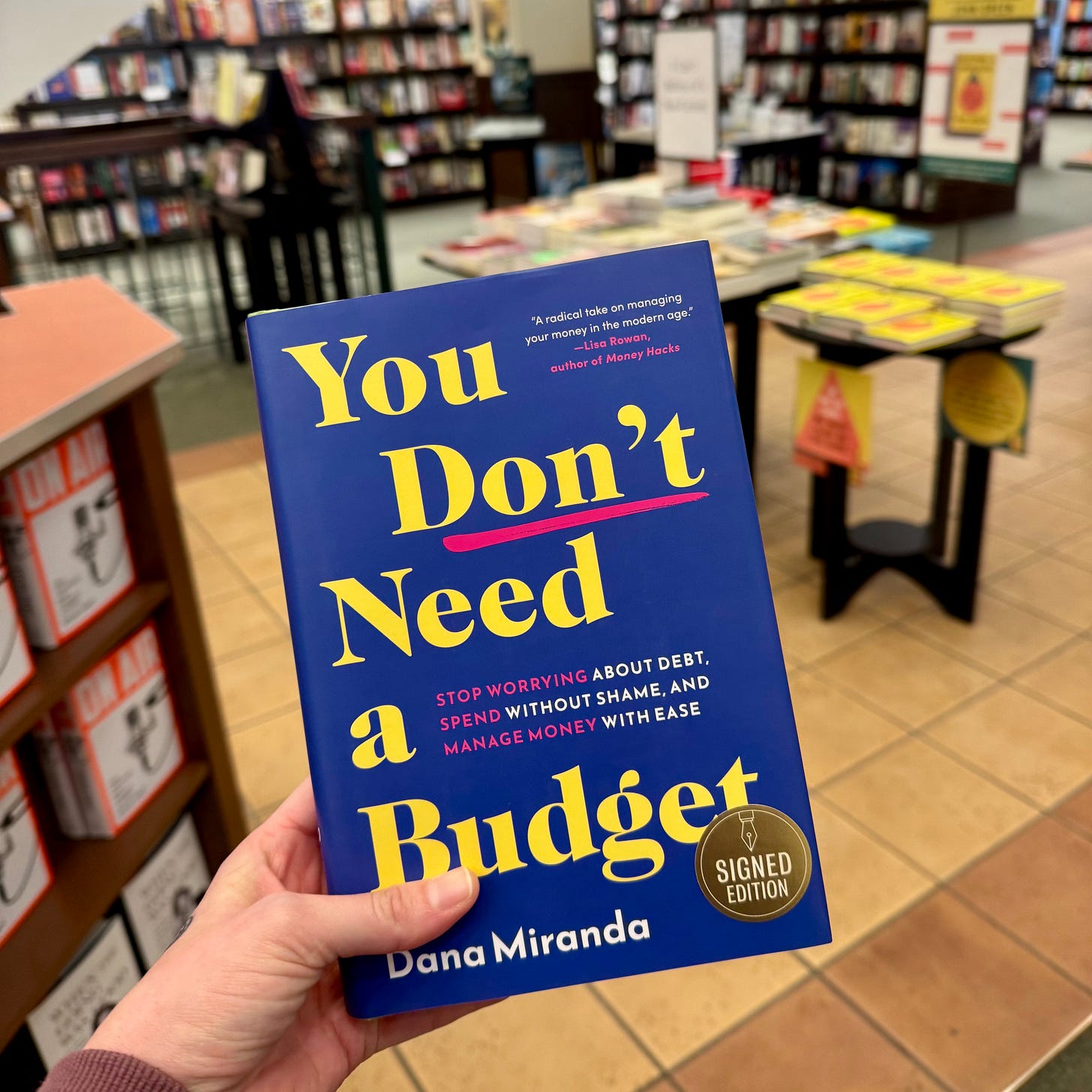 Photo of a hand holding a hardcover copy of YOU DON'T NEED A BUDGET by Dana Miranda, with a "signed copy" sticker on the front, in front of shelves of books.
