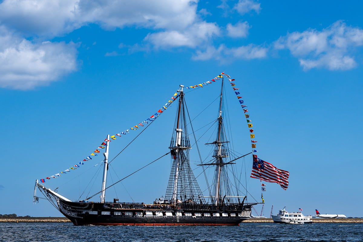 USS Constitution sailing Boston Harbor with flags and tug escort near Seaport skyline USS Constitution sailing Boston Harbor with flags and tug escort near Seaport skyline