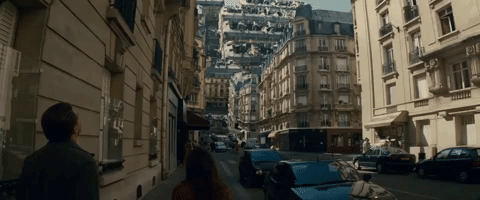 Gif con l'nquadratura dal film di fantascienza "Inception" che mostra una strada cittadina in una città europea (probabilmente Parigi) con edifici in stile haussmanniano ai lati. L'elemento surreale è dato dal fatto che in lontananza, le facciate degli edifici sembrano piegarsi e ripiegarsi su sé stesse, sfidando la gravità e creando una vertiginosa struttura a specchio. Due figure sfocate, apparentemente un uomo e una donna, camminano in primo piano dando le spalle all'osservatore.