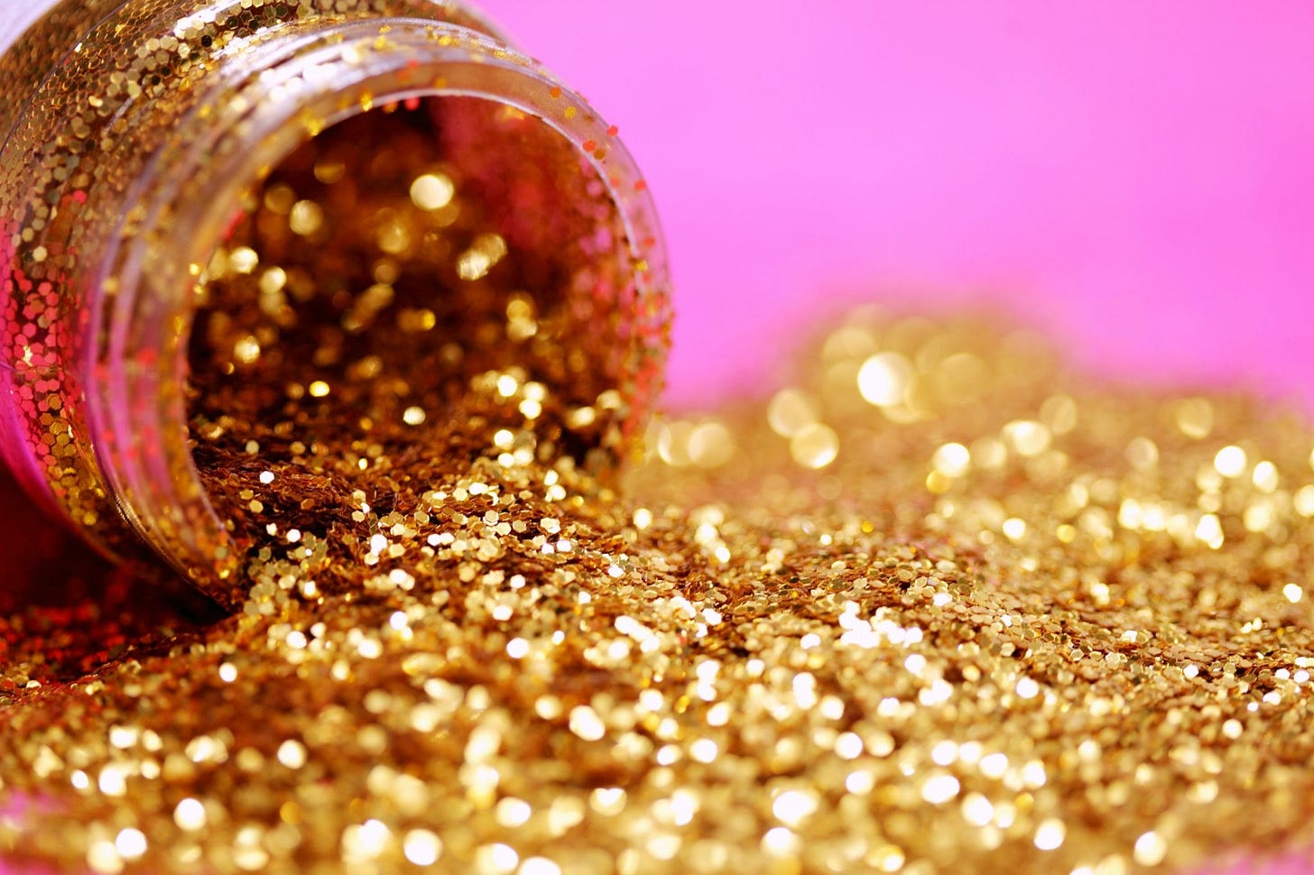 Close-up of gold glitter spilling from a small jar onto a surface, with soft pink and gold light creating a blurred, shimmering background.