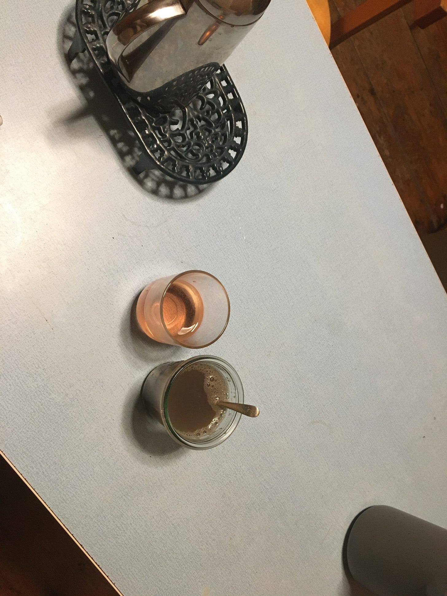 A shot of a blue formica table at a zany angle. It is night and the overhead light’s harsh bright is reflected in a spot on the table. At the top, cut off, is a green ornate metal trivet with a silver coffee pot on it. At the bottom, also cut off, is a grey jug og water. Just off centre are two glasses, one, higher up, with a pink clear liquid in, the other with a murky green liquid in, and a spoon. 