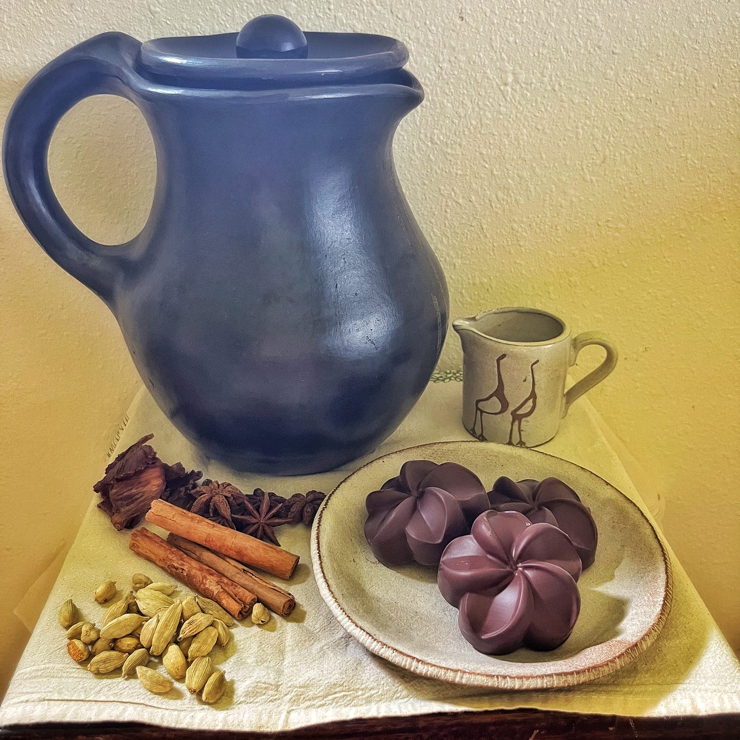 Still life photo with black clay pitcher, pottery creamer, spices, and chocolate molded into a plumeria shape