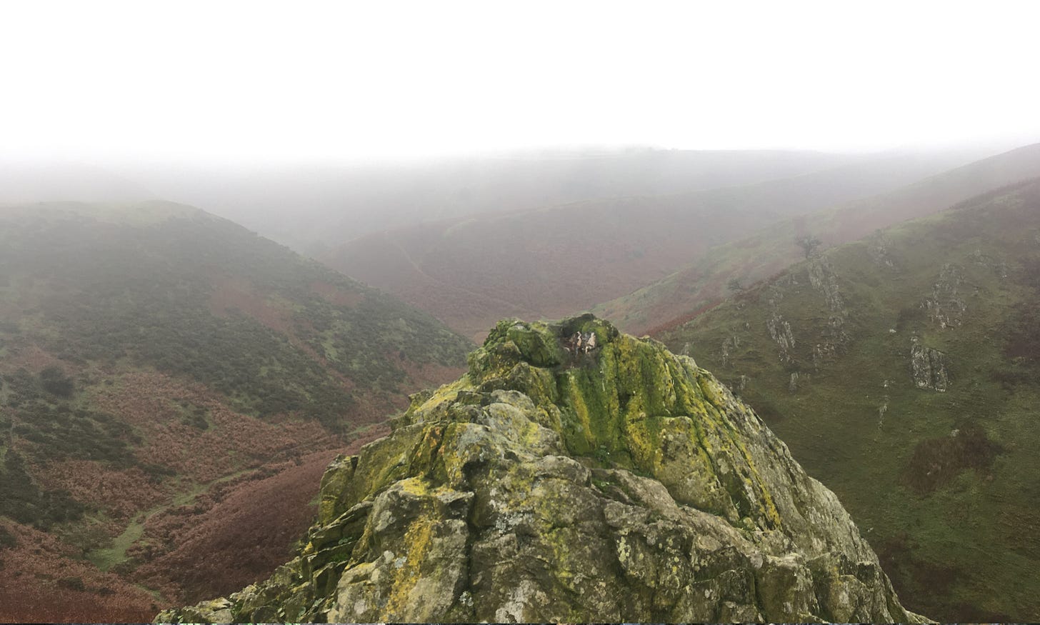 in the foreground is a rock, with green moss / lichen on, with a view over hills and a valley of green and brown grass and bracken. with a grey white foggy sky above. in the foreground is a rock, with green moss / lichen on, with a view over hills and a valley of green and brown grass and bracken. with a grey white foggy sky above.