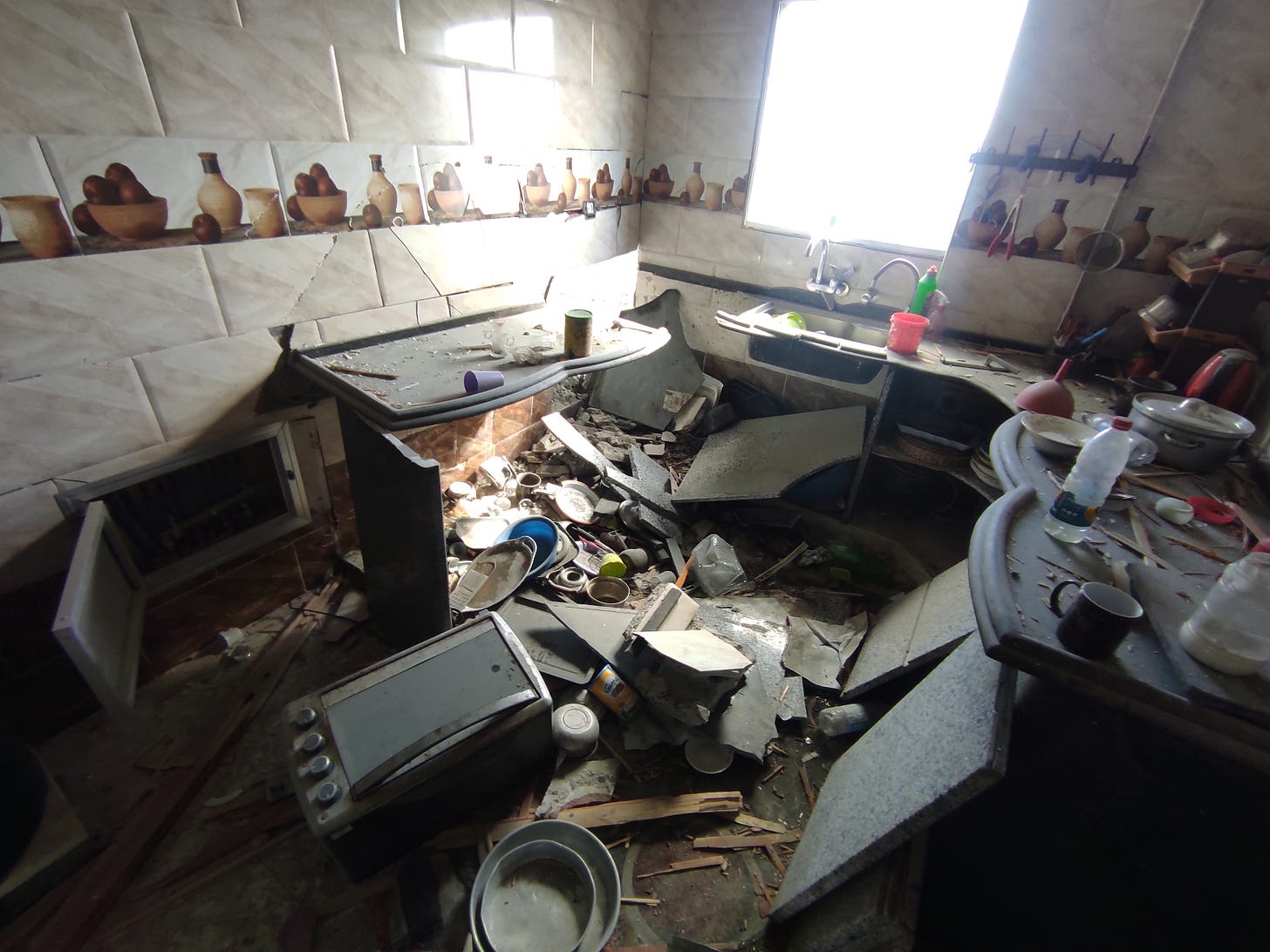 A heavily damaged kitchen filled with rubble and debris. Several dark stone countertops have collapsed into a large hole or sunken area in the floor at the center of the room. Broken countertop slabs, shattered tiles, pieces of wood, dishes, pots, and kitchen utensils are scattered across the floor. A microwave lies tipped over in the foreground, and cabinet doors hang open. Bright sunlight streams through a window above a double sink, illuminating the destruction. Decorative wall tiles featuring pottery and fruit remain intact along the walls, contrasting with the extensive structural damage below. A heavily damaged kitchen filled with rubble and debris. Several dark stone countertops have collapsed into a large hole or sunken area in the floor at the center of the room. Broken countertop slabs, shattered tiles, pieces of wood, dishes, pots, and kitchen utensils are scattered across the floor. A microwave lies tipped over in the foreground, and cabinet doors hang open. Bright sunlight streams through a window above a double sink, illuminating the destruction. Decorative wall tiles featuring pottery and fruit remain intact along the walls, contrasting with the extensive structural damage below.