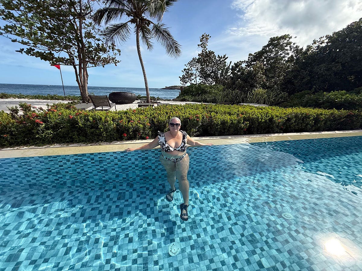 Writer Kristi Koeter standing in the Six Senses La Sagesse Grenada resort pool with ocean and palm trees in the background