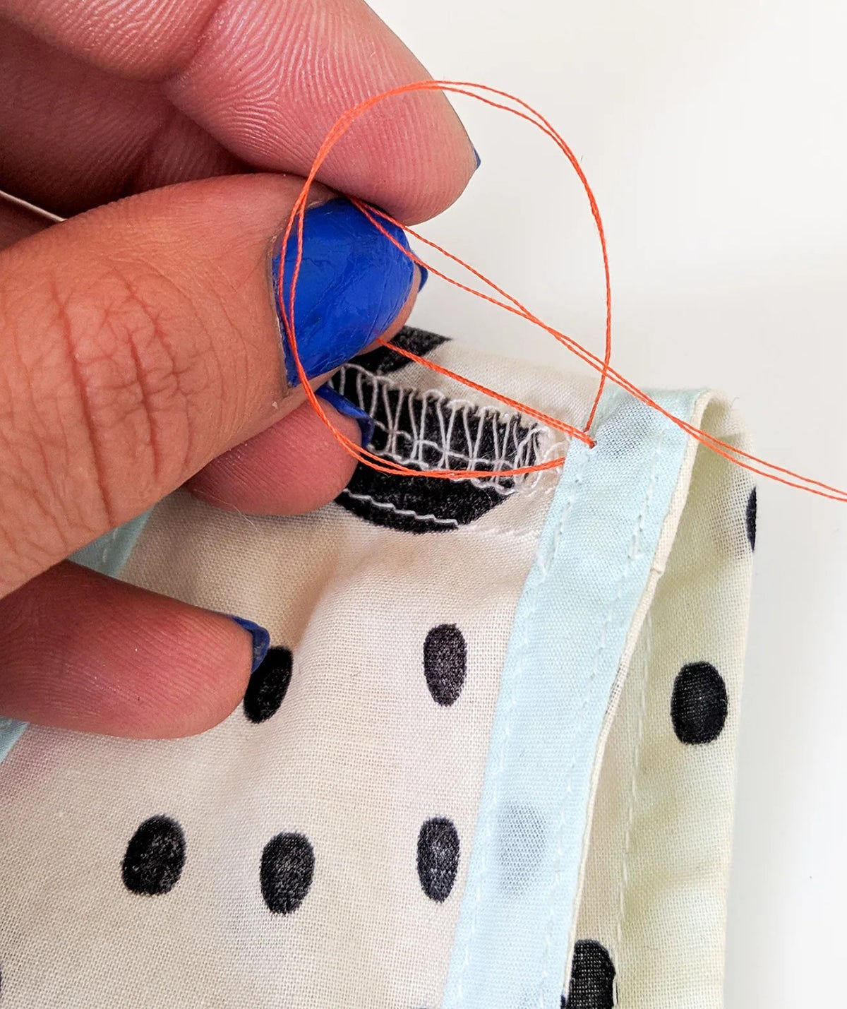 Needle pulling orange thread through fabric near armhole seam with serged edge and light blue binding.