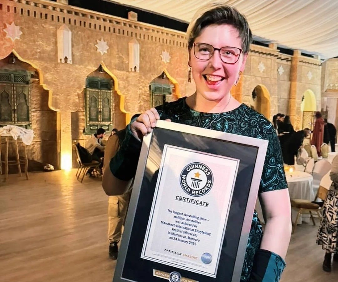 Iris Skipworth, a white woman with short blinde hair, red glasses, and a green dress and gloves, stands smiling at the camera holding a framed certificate. The certificate has a silver frame and says 'Guinness World Records Certificate: The longest storytelling show - multiple storytellers - was achieved by Marrakech International Storytelling Festival (Morocco) in Marrakech Morocco on 24 January 2025'. She is standing in a room of tables and chairs, with stars on the wall and a cloth ceiling, lit up by lamps.