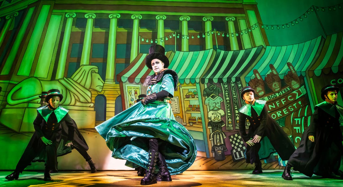 A hand-painted backdrop of Nottingham’s Market Square, featuring the Left Lion and various market stalls. Centre stage, lit by a spotlight, is a man wearing a top hat, green long cloak, and black knee-high boots. He is swishing his cloak. Four young people stand either side of him, wearing black hats and black cloaks with green collars. They are leaning their weight to one side, with their hands on their knees.