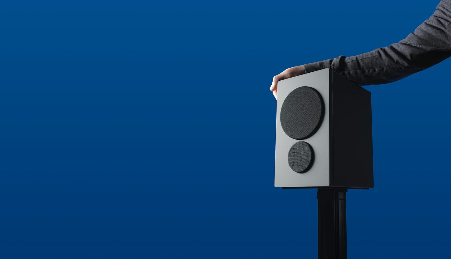 Someone's hand sits atop a PSB iQ speaker mounted on a floor stand against a blue background.