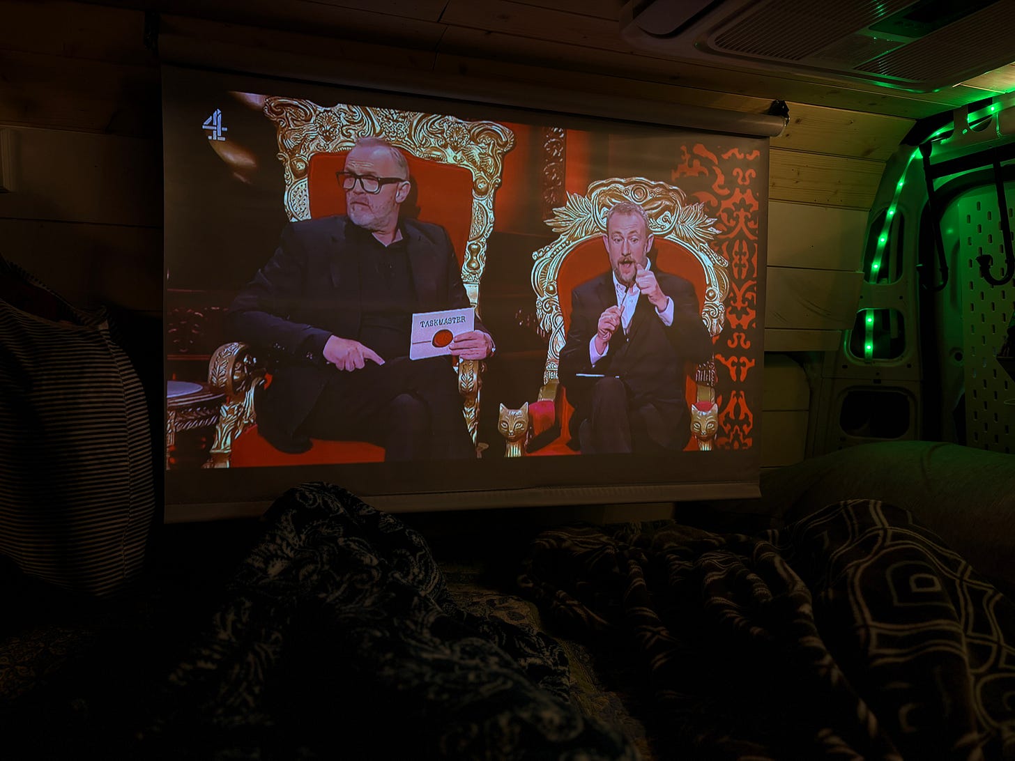 A projector screen inside a van shows a scene from the TV show Taskmaster with Greg Davies and Alex Horne sitting on ornate golden thrones. The shot is taken from inside the van’s bed area, with blankets visible in the foreground and warm interior lighting adding to the cozy atmosphere.