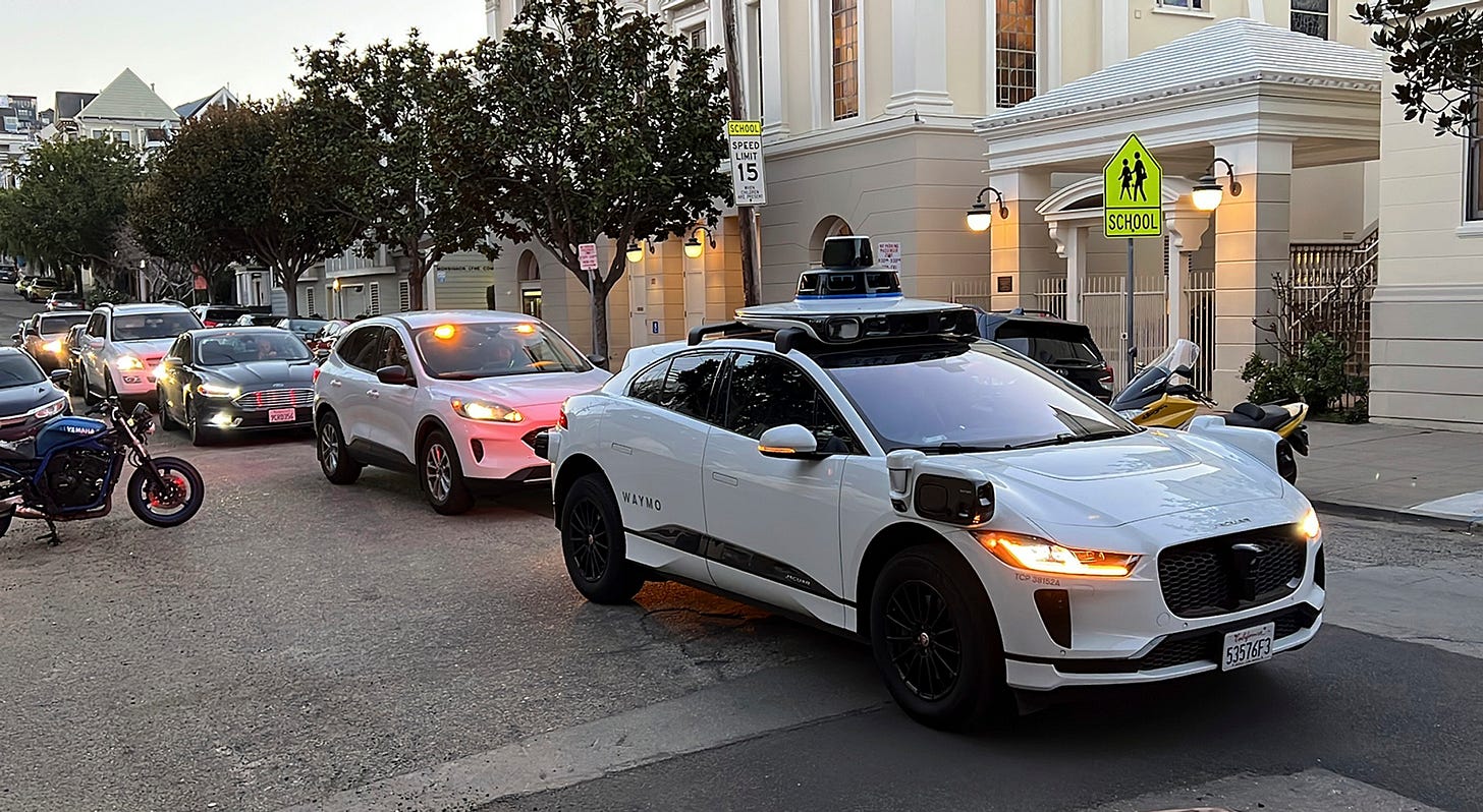 A white car with running lights on and with the word \u201cWaymo\u201d emblazoned on the rear door stands in a street, with other cars backed up behind it.