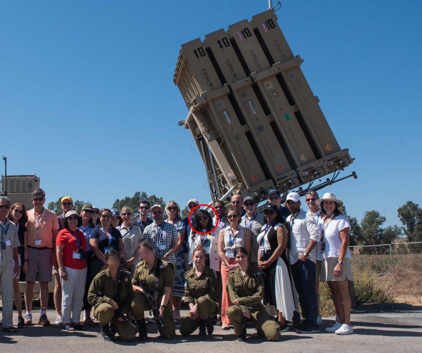 A group of roughly two dozen civilians, some wearing conference lanyards, pose in front of an Iron Dome missile defense battery under a clear blue sky. Three IDF soldiers crouch in the foreground. Rep. Jasmine Crockett is circled in red.