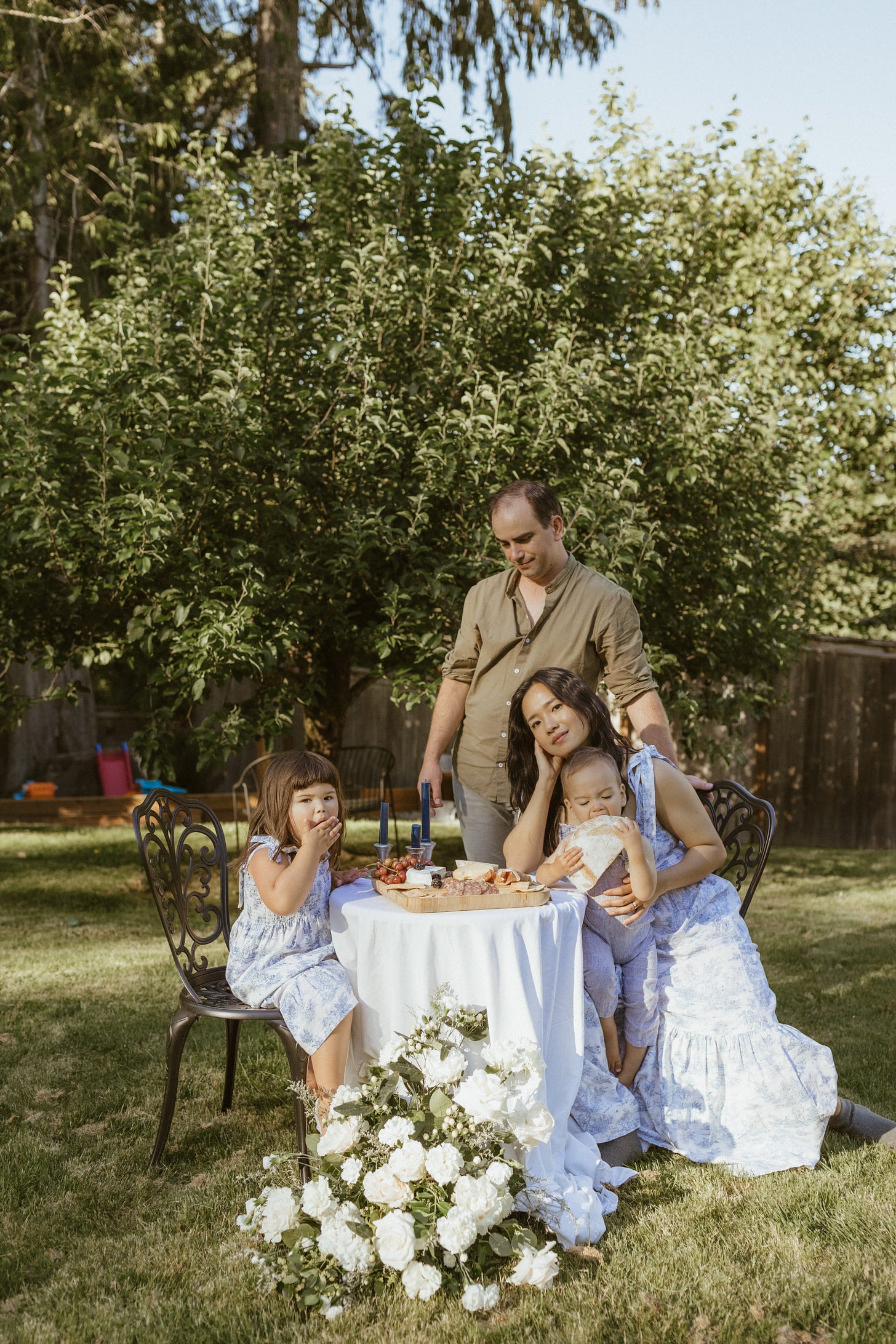 A family of four sits around a decorated picnic table outdoors, all wearing white outfits