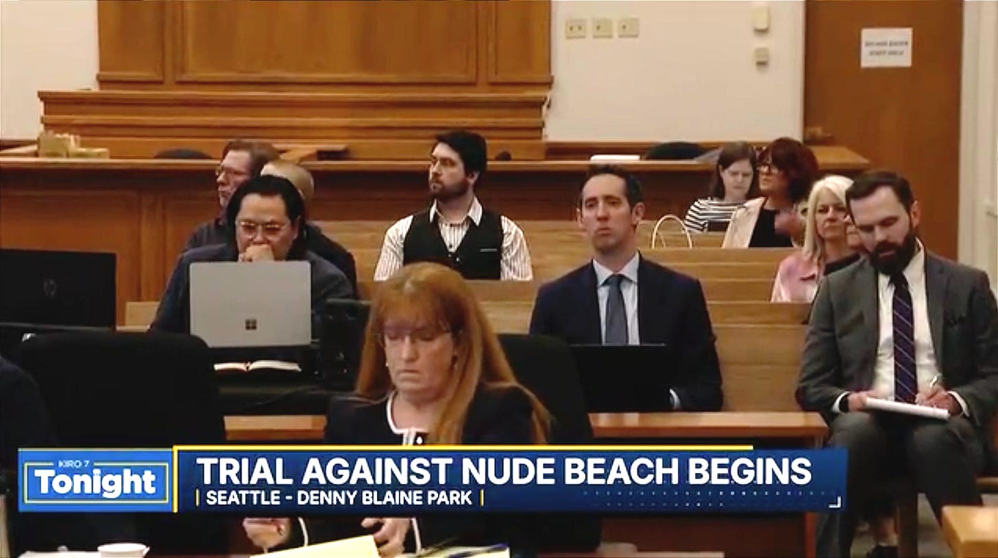 Alt text: A KIRO 7 news still from inside a Seattle courtroom shows rows of people seated in wooden gallery benches. In the front row, a woman with red hair in a dark blazer looks down at papers on the table before her. Behind her sit several observers, including a man in a black vest over a pinstriped shirt and knit cap, a clean-cut man in a navy suit and tie, and a bearded man in a gray suit writing on a notepad at right. A chyron reads "TRIAL AGAINST NUDE BEACH BEGINS" with the subhead "SEATTLE — DENNY BLAINE PARK." Alt text: A KIRO 7 news still from inside a Seattle courtroom shows rows of people seated in wooden gallery benches. In the front row, a woman with red hair in a dark blazer looks down at papers on the table before her. Behind her sit several observers, including a man in a black vest over a pinstriped shirt and knit cap, a clean-cut man in a navy suit and tie, and a bearded man in a gray suit writing on a notepad at right. A chyron reads "TRIAL AGAINST NUDE BEACH BEGINS" with the subhead "SEATTLE — DENNY BLAINE PARK."