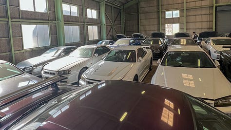 A collection of photos showing dozens of white, blue and green Nissan R34 Skyline GT-R sports cars crammed into a dimly-lit warehouse with gray walls and green support beams with a gravel floor.