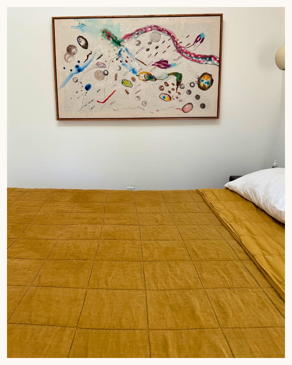 Wide bedroom view with mustard quilt and a horizontal mixed-media textile work by Vanessa Valero (2024) in a slim wood frame above the bed.