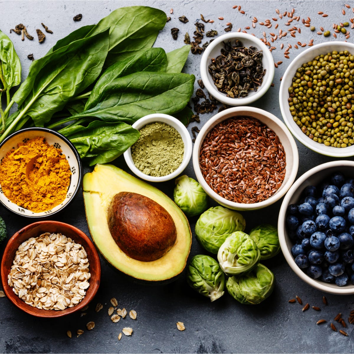 healthy foods including fresh spinach, avocado, oats, flaxseeds, mung beans, blueberries, Brussels sprouts, turmeric, and green tea on a grey surface healthy foods including fresh spinach, avocado, oats, flaxseeds, mung beans, blueberries, Brussels sprouts, turmeric, and green tea on a grey surface