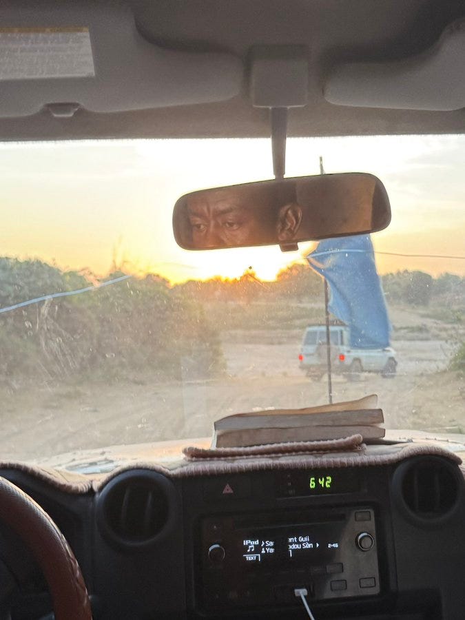 View from inside a vehicles dashboard showing a rearview mirror reflecting the drivers face partially, a book on the dashboard, digital display showing time and fuel gauge, steering wheel, and through the windshield a dirt road at sunset with sparse vegetation, a white vehicle ahead, utility poles, and orange sky.