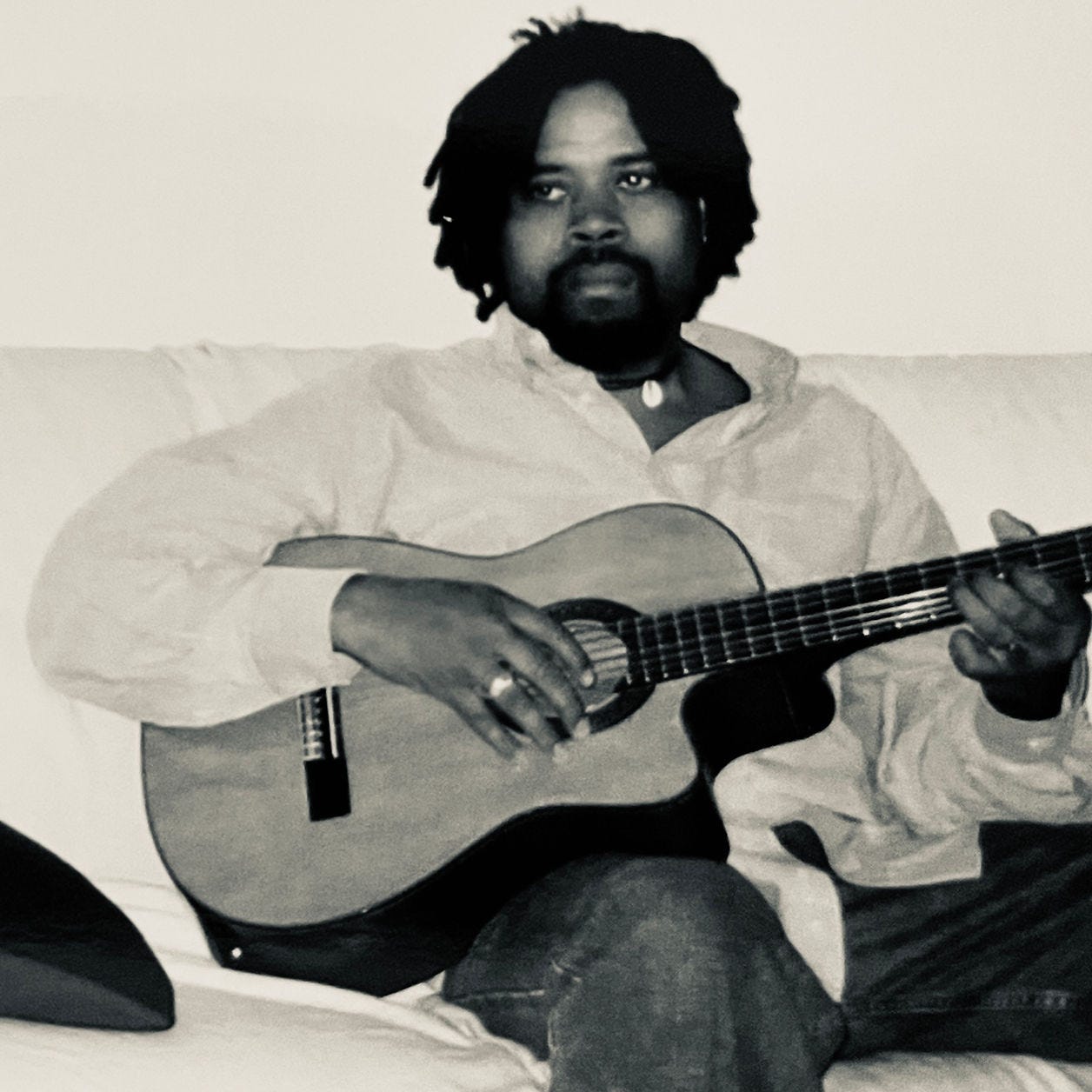 A man with locks plays an acoustic guitar on a white couch, wearing a light shirt. The mood is calm and introspective. Black and white image.