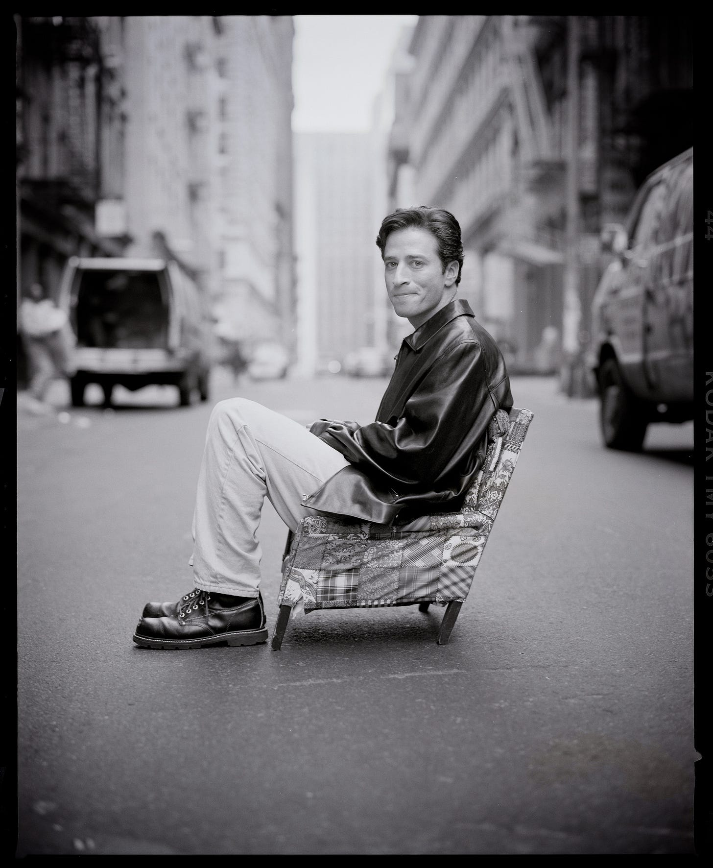 Jon Stewart sitting in a tiny chair in the middle of the street.