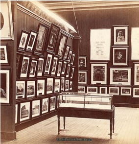 Interior view of an art gallery featuring a wall covered with framed photographs and art pieces, with a display case in the center.