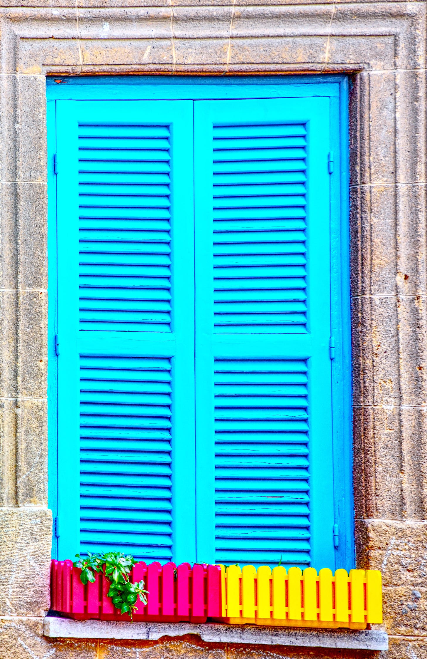 Blue shutters on a window in Kyrenia, northern Cyprus, photographed by Jay Siegmann, featuring a colorful flower box with red, yellow, and green accents.