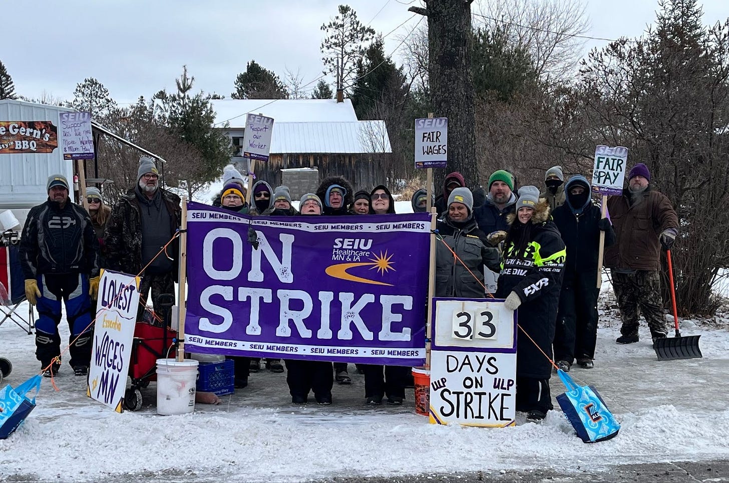 a dozen or so people stand on a snowy sidewalk wearing winter gear and holding picket signs and a purple banner that reads "on strike" a dozen or so people stand on a snowy sidewalk wearing winter gear and holding picket signs and a purple banner that reads "on strike"