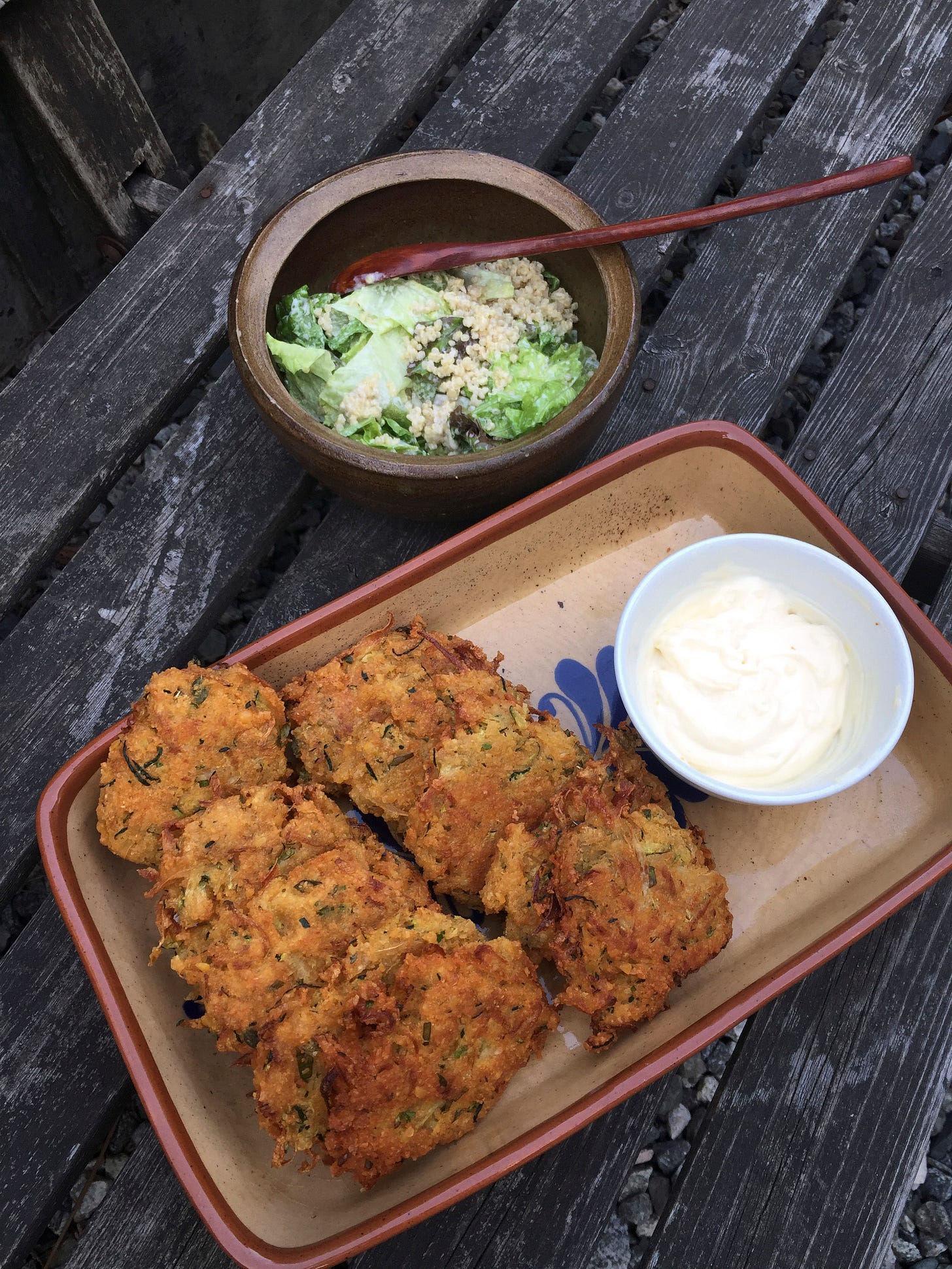 On a wooden bench, a rectangular dish full of golden-brown fritters, next to a small white dish of the lemon mayo. Behind it is a small stone bowl of quinoa Caesar salad. On a wooden bench, a rectangular dish full of golden-brown fritters, next to a small white dish of the lemon mayo. Behind it is a small stone bowl of quinoa Caesar salad.