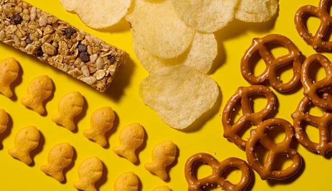 Lay's Classic Potato Chips, Quaker Chewy Bars Chocolate Chip, Snyder's of Hanover Mini Pretzels, and Pepperidge Farm Goldfish Cheddar Crackers arranged on a bright yellow background. Lay's Classic Potato Chips, Quaker Chewy Bars Chocolate Chip, Snyder's of Hanover Mini Pretzels, and Pepperidge Farm Goldfish Cheddar Crackers arranged on a bright yellow background.