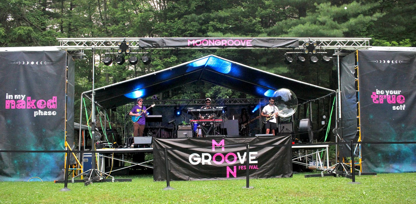 Hot Like Mars performing on MoonGroove's outdoor main stage, four musicians playing guitar, keyboard, and bass under string lights and a tented rig, with MoonGroove festival signage in the foreground. Hot Like Mars performing on MoonGroove's outdoor main stage, four musicians playing guitar, keyboard, and bass under string lights and a tented rig, with MoonGroove festival signage in the foreground.