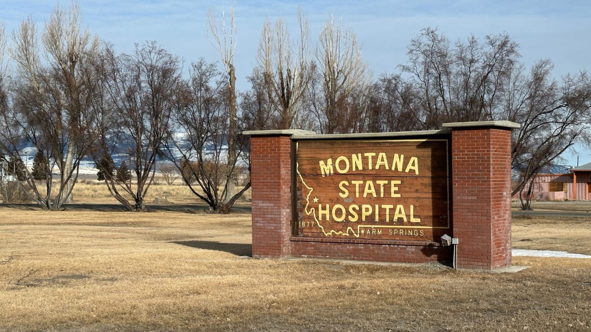 A brick sign with the inscription "Montana State Hospital - 1877 Warm Springs" stands in a grassy area with leafless trees in the background. In the distance, snow-covered mountains can be seen under a clear sky.