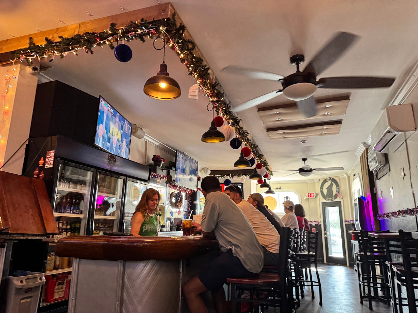 A scene, showing a bartender and people sitting at the bar. There's a television showing FIFA on the wall behind the bar, and a ceiling fan above. 