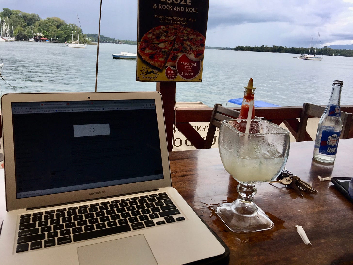 A laptop sits on a table with a margarita next to it overlooking the water.
