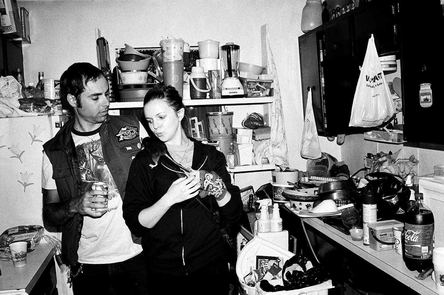 Derrick and Amber stand together in a small, cluttered kitchen. There is a rat on Amber's shoulder. The choker she appeared to be wearing was actually the rat's tail. Both Derrick and Amber look down at the rat as it climbs on Amber.