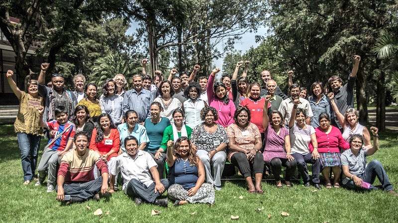 Group photo of 30+ people, on a sunny day, sitting on green grass with trees behind them. There is a front row of people sitting down and multiple rows of people standing behind them. It is mostly women, with some men, mostly Black and Brown skinned people. Some are raising their fists. Lots are smiling.