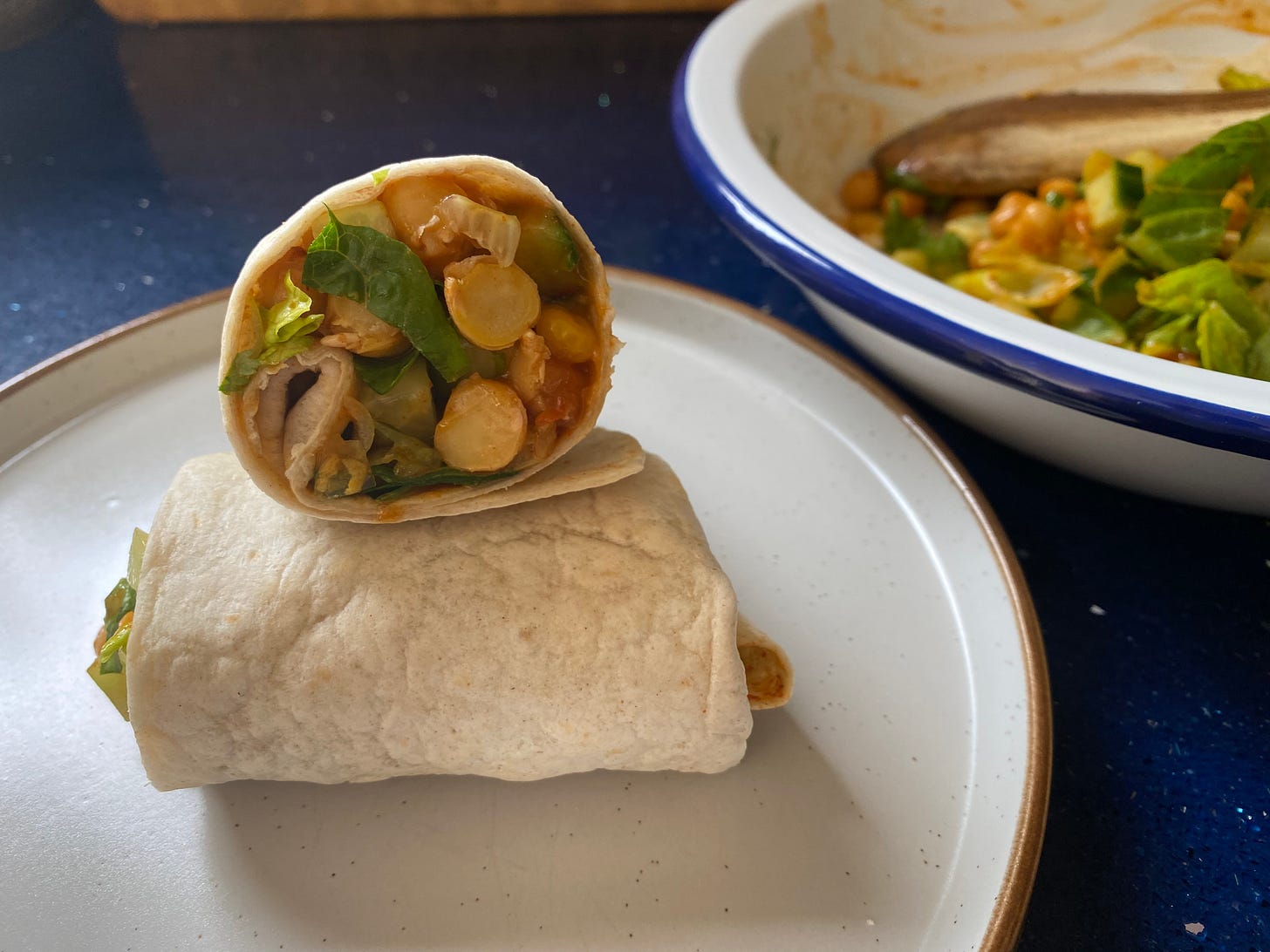 Cut open side of a tortilla wrap sandwich showing chickpeas and salad. A blue and white bowl of the sandwich filling is placed to the right.