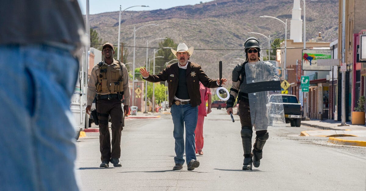 Phoenix stands in front of a street protest holding a ring light, flanked by two police officers in tactical gear addressing demonstrators. Phoenix stands in front of a street protest holding a ring light, flanked by two police officers in tactical gear addressing demonstrators.