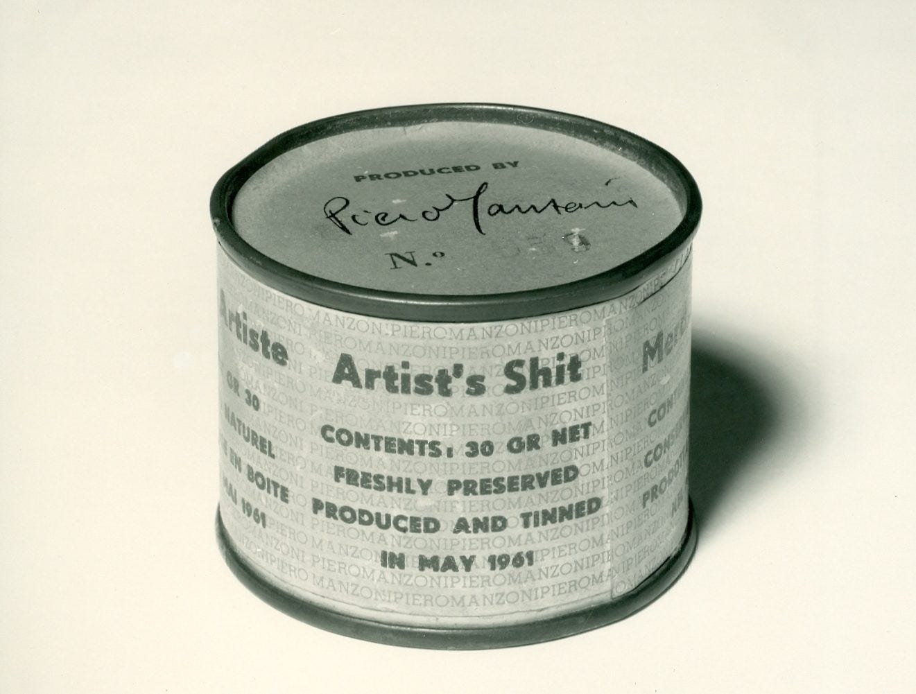 Artist’s Shit by Piero Manzoni – a grey tin with a paper label. The paper label has a grey text background of the repeated word PIEROMANZONI, over which, in several languages, appear the words: Artist’s Shit. Contents: 30 gr. Net, Freshly preserved. Produced and tinned in May 1961. In this image, the English version of the text is to the front