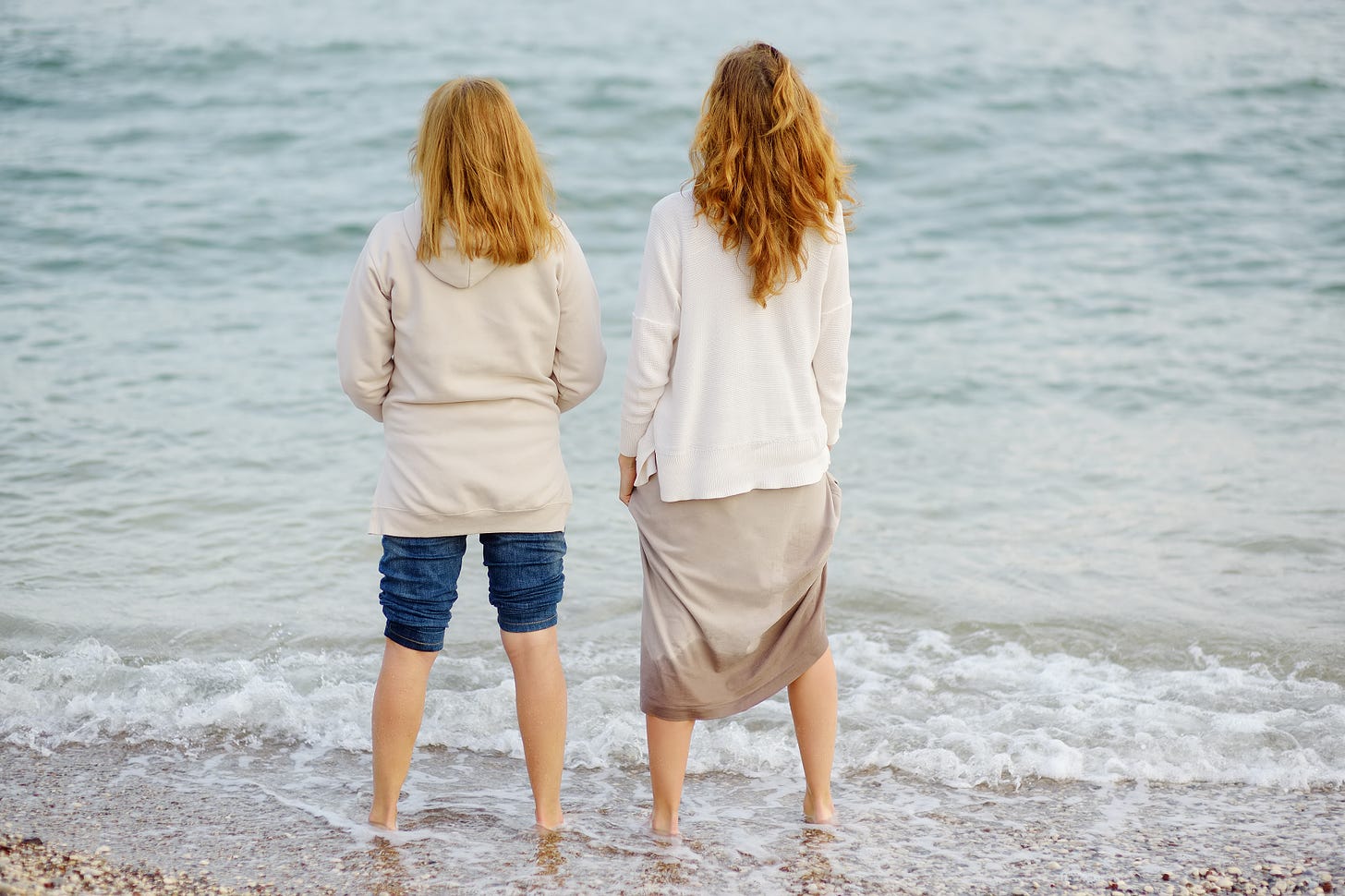 An older woman and a younger woman stand side by side at the water’s edge, their backs to the camera. Gentle waves lap around their feet as they look out over a calm sea.