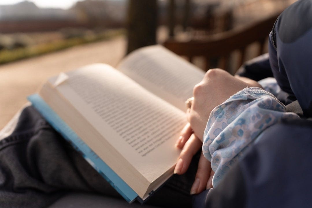 Person reading an open book outdoors on a bench.