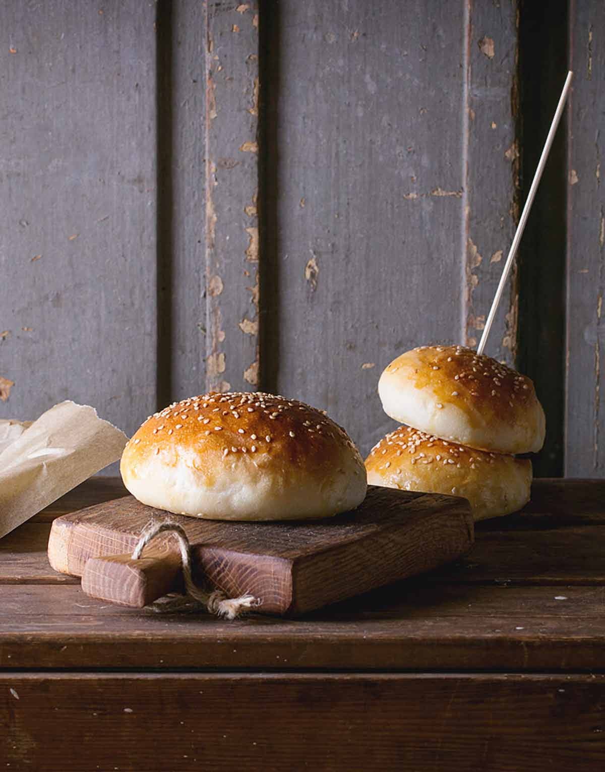 Three golden brown brioche hamburger buns topped with sesame seeds on a cutting board.