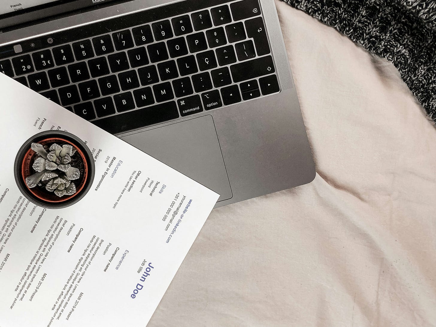 A silver laptop with a black keyboard sits on a white blanket. To the left covering part of the keyboard is a white resume with black letters in small unintelligible font. A black cup sits on top of the resume, filled with something gray.