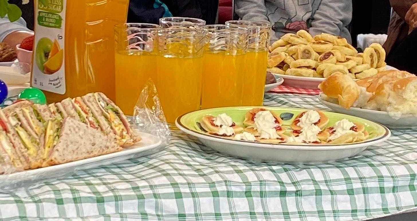 table spread with sandwiches, glasses of juice, sausage rolls, and pikelets